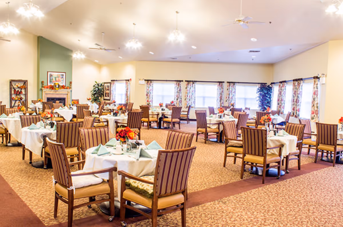 A spacious dining room with multiple round tables covered with white tablecloths, each set with folded napkins, cups, and floral centerpieces. The room has patterned carpet, large windows with floral curtains, ceiling fans, and warm lighting. There is a fireplace with decorations on the far wall.