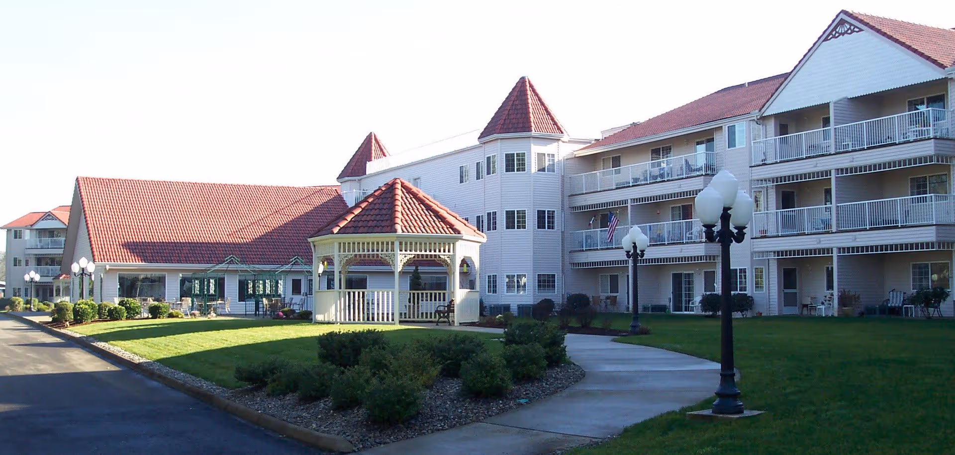 Exterior view of Riverview Terrace senior living facility showing a multi-story building with balconies, a gazebo with a red roof in the foreground, well-maintained green lawn, shrubs, and lamp posts along a paved walkway.