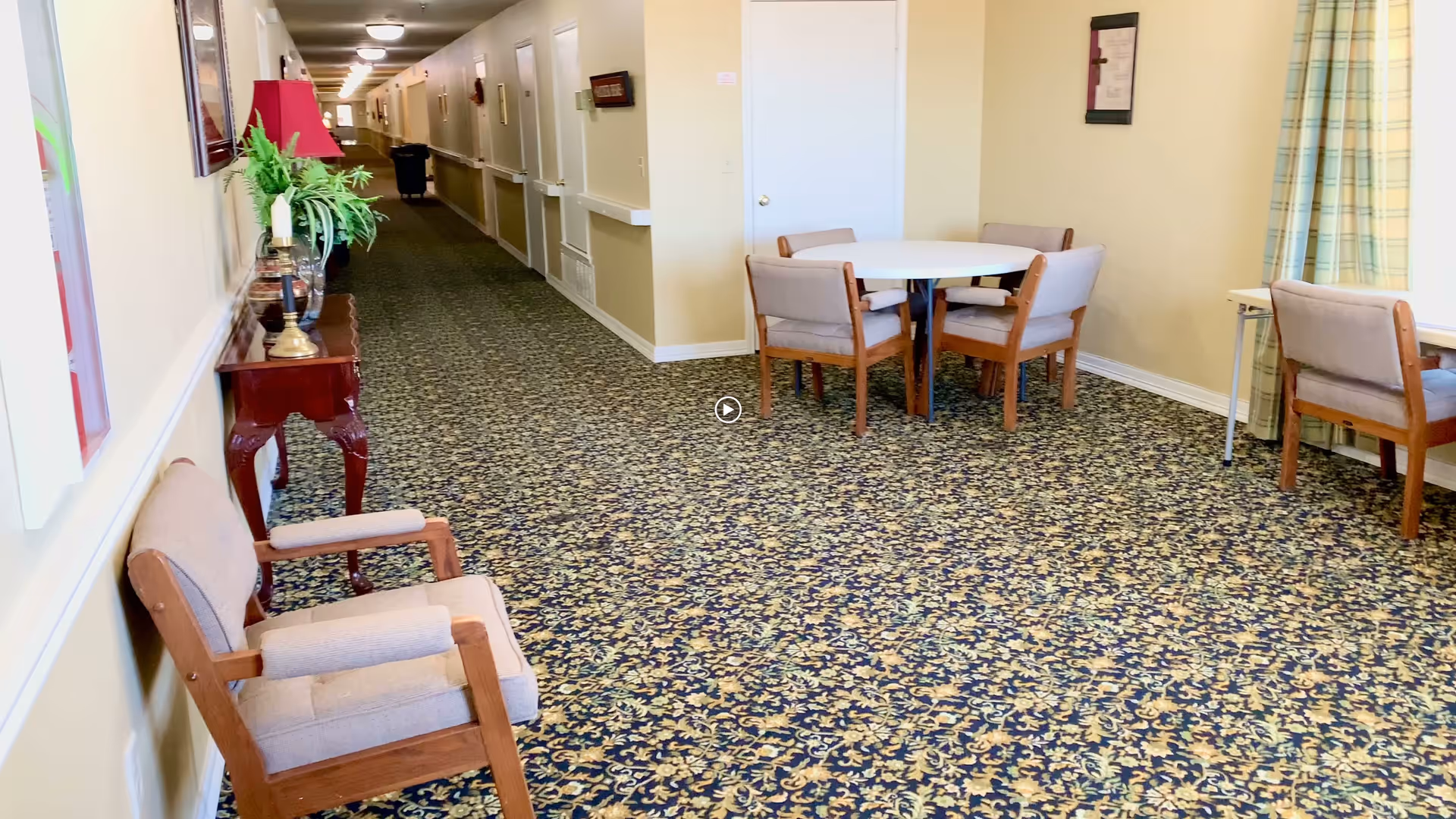 A long hallway in a senior living facility with patterned carpet, beige walls, and several doors along the corridor. On the left side, there is a wooden table with a red lamp and a plant. On the right side, there is a small seating area with a round table and four chairs, as well as another chair near a window with plaid curtains.
