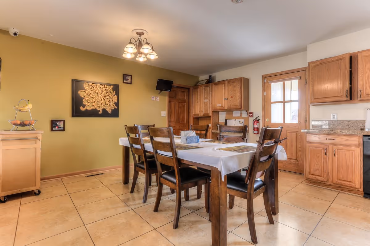 A dining area with a wooden table covered with a white tablecloth and six wooden chairs with black cushions. The room has tiled floors, wooden cabinets, a wooden door with glass panels, and a green accent wall with decorative artwork. A ceiling light fixture with multiple bulbs hangs above the table.