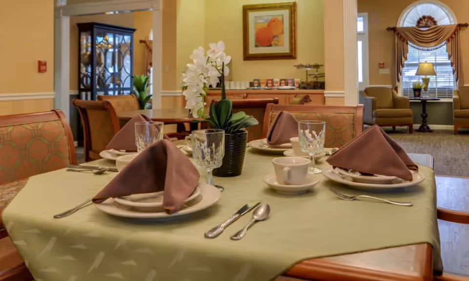 A dining table set for four with brown folded napkins, white plates, cups, and crystal glasses on a light green tablecloth. In the background, there is a cabinet with glass doors, a painting on the wall, and a seating area with armchairs and a window with drapes.