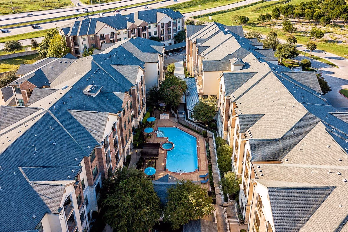 Aerial view of a senior living facility named Conservatory At North Austin showing multiple connected buildings surrounding a central courtyard with a swimming pool, hot tub, umbrellas, and seating areas. Trees and greenery are visible around the courtyard and along the perimeter of the buildings. A highway and parking areas are visible in the background.
