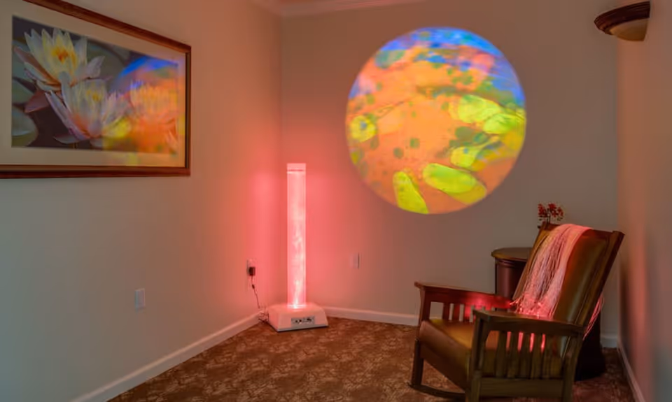 A small sitting area with a wooden armchair draped with a throw, a tall illuminated sensory lamp in the corner, and a colorful circular light projection on the wall.