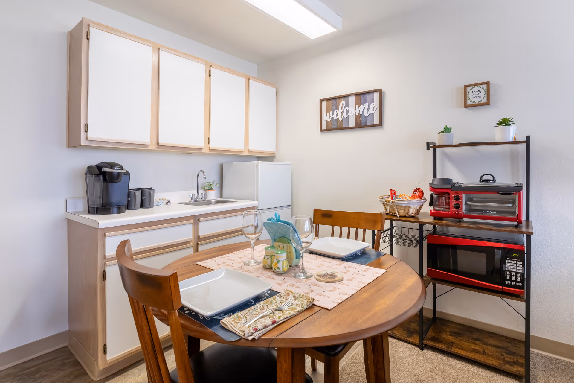 A small kitchen and dining area with a round wooden table set for two with plates, glasses, and napkins. The kitchen has white cabinets with light wood trim, a small sink, a coffee maker, and a mini refrigerator. A shelving unit holds a red microwave, toaster oven, coffee maker, and a basket with snacks. Wall decor includes a wooden 'welcome' sign and a small framed sign that says 'Home Sweet Home'.