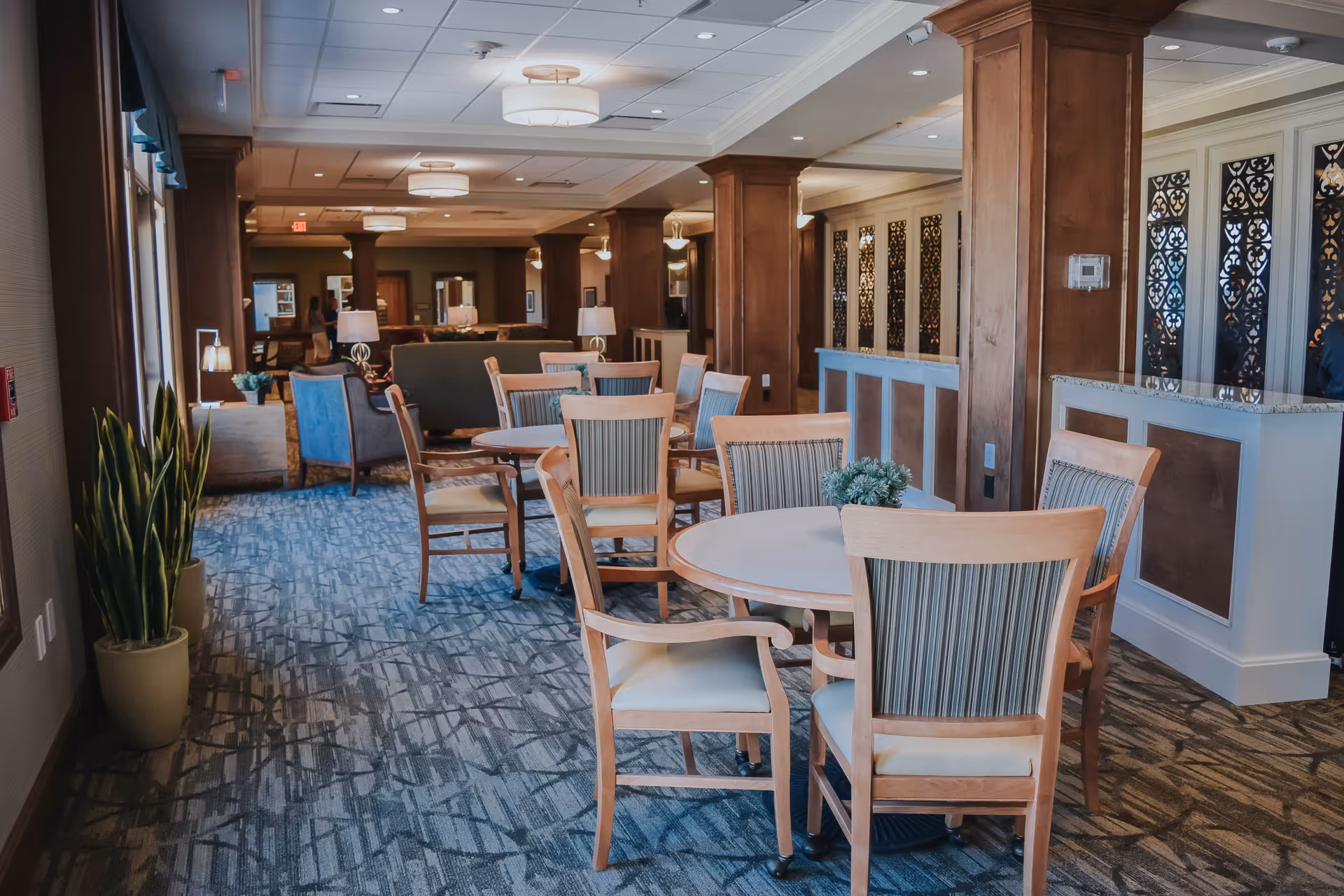 Interior view of a senior living facility common area with round tables and wooden chairs arranged on a patterned carpet. The room features wooden columns, decorative wall panels, ceiling lights, and potted plants near the windows.