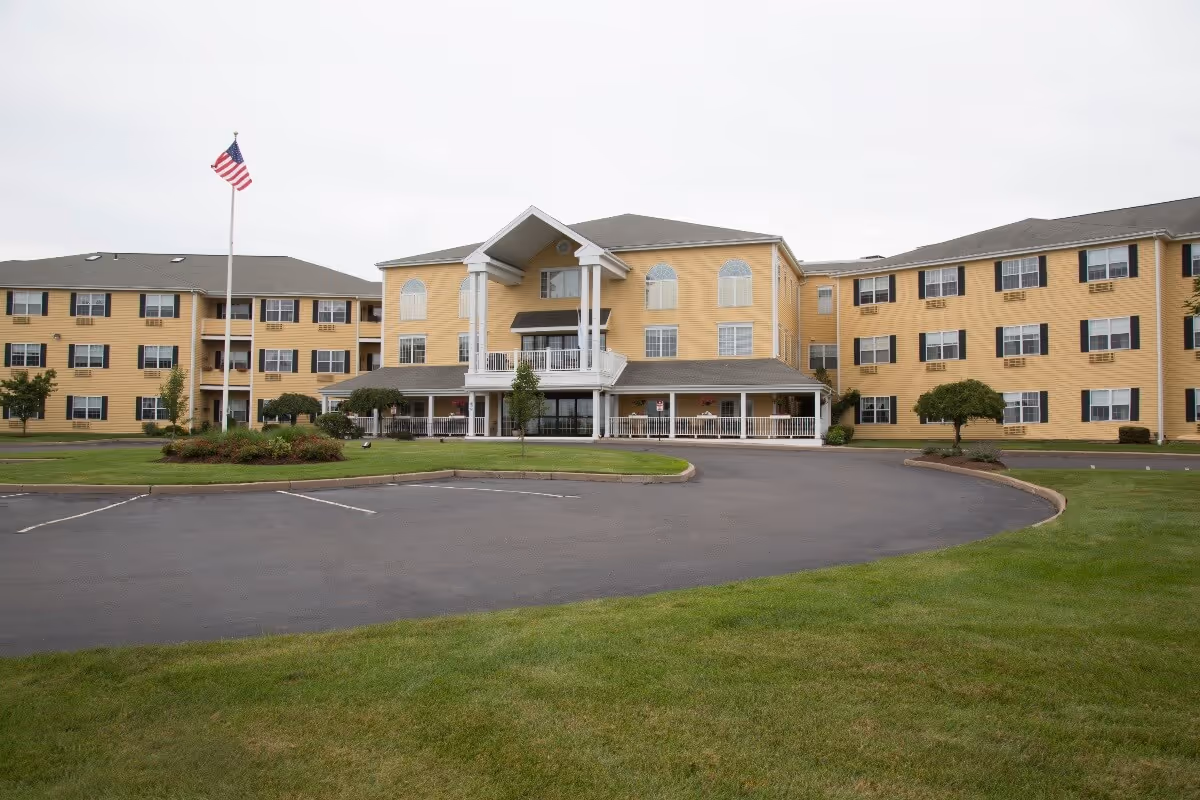 Exterior view of a large, three-story yellow senior living facility building with white trim and black shutters. The building has a covered entrance with white columns and a balcony above it. There is a circular driveway and a flagpole with an American flag in front, surrounded by green grass and small trees.
