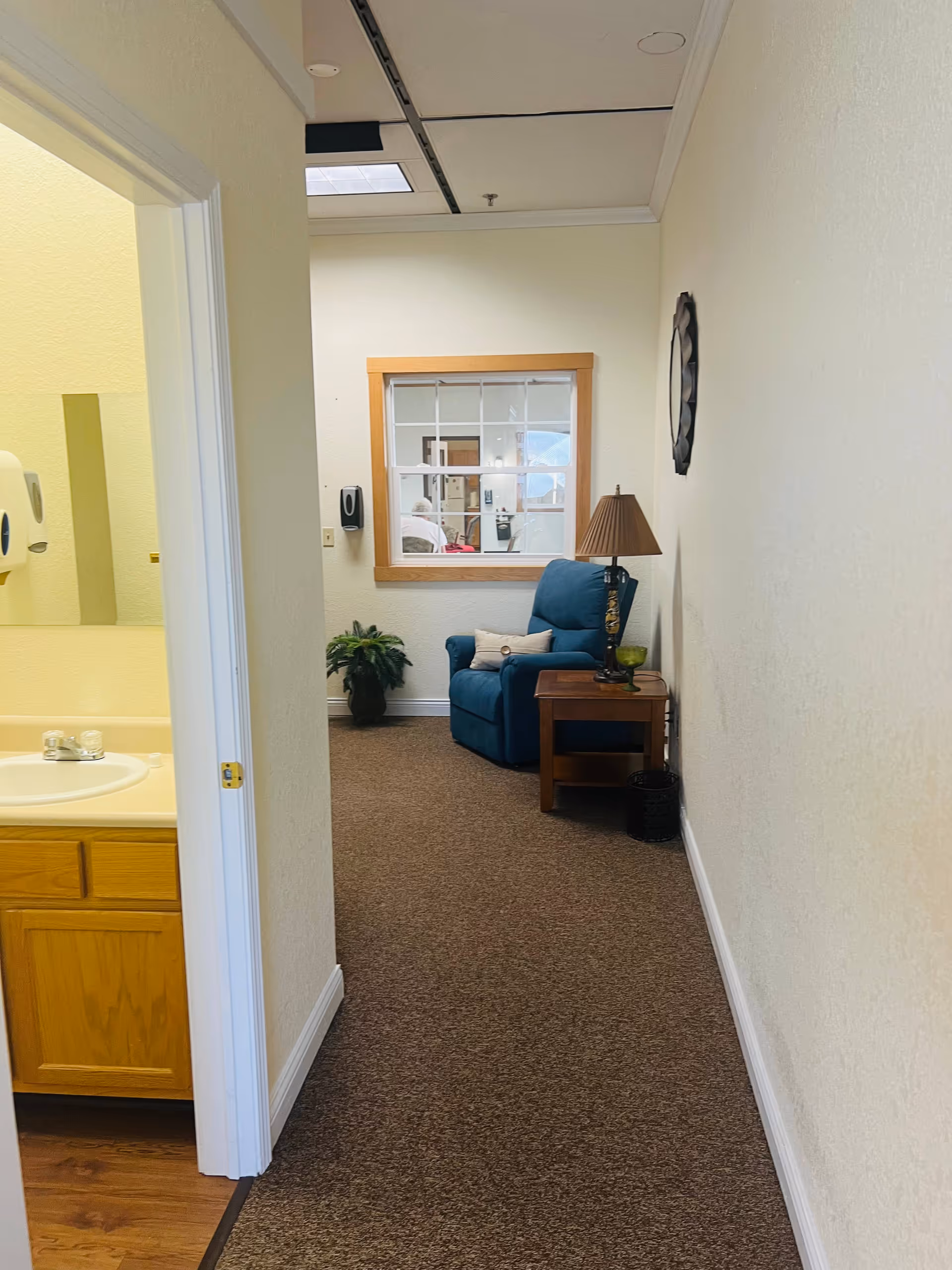 A hallway with beige walls and brown carpet leading to a blue armchair next to a wooden side table with a lamp. A window with wooden trim is on the far wall, and a plant is placed near the window. To the left, there is a partial view of a bathroom sink with a wooden cabinet and a soap dispenser on the wall.