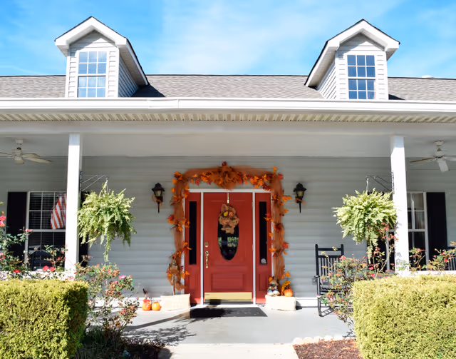 Front porch of a building with a red door decorated for autumn, flanked by hanging ferns, rocking chairs, and trimmed shrubs.