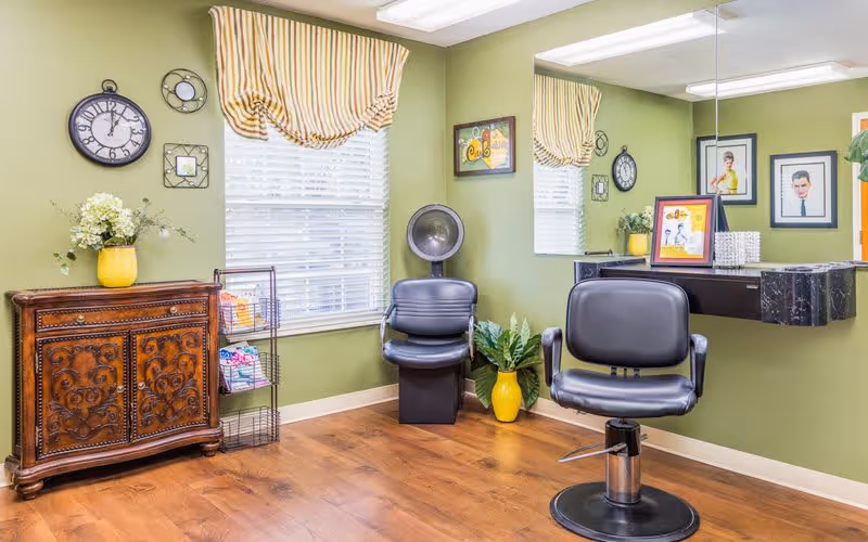 A small salon area with two black salon chairs, one in front of a wall-mounted mirror and the other under a vintage hair dryer. The room has green walls, a wooden floor, a window with striped valance curtains, a decorative wooden cabinet with a yellow vase of flowers, and framed pictures on the walls.