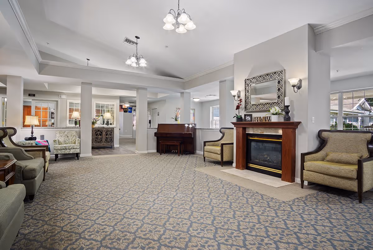 A spacious and well-lit living room area in a senior living facility with patterned carpet flooring, multiple armchairs, a wooden fireplace with decorative items and a mirror above it, a piano against the wall, and several lamps providing warm lighting.