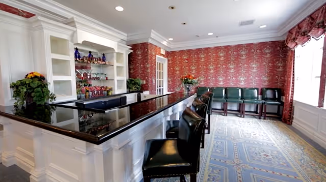 Interior view of a room with a long black countertop bar with several black leather bar stools. The room features white cabinetry with shelves displaying decorative items, red patterned wallpaper, a large window with red curtains, and a carpeted floor with a blue and beige design. There are additional green chairs lined up against the wall near the window.