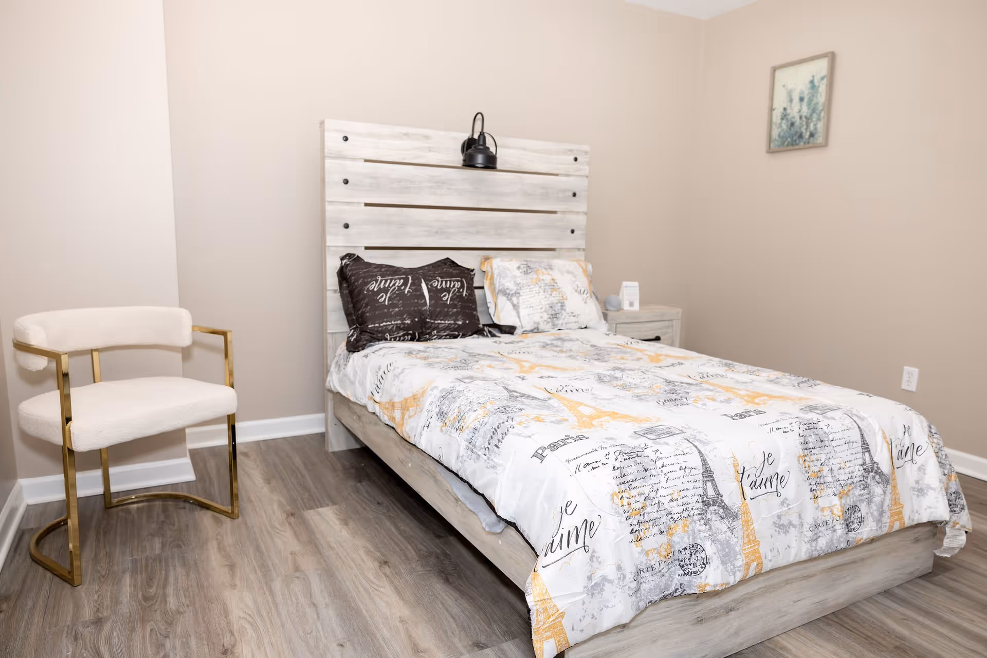 A bedroom with a wooden bed frame and headboard, a bed covered with a white and yellow patterned comforter featuring Eiffel Tower and script designs, two pillows, a small nightstand with a small item on it, a modern white and gold chair, light wood flooring, beige walls, and a framed picture hanging on the wall.