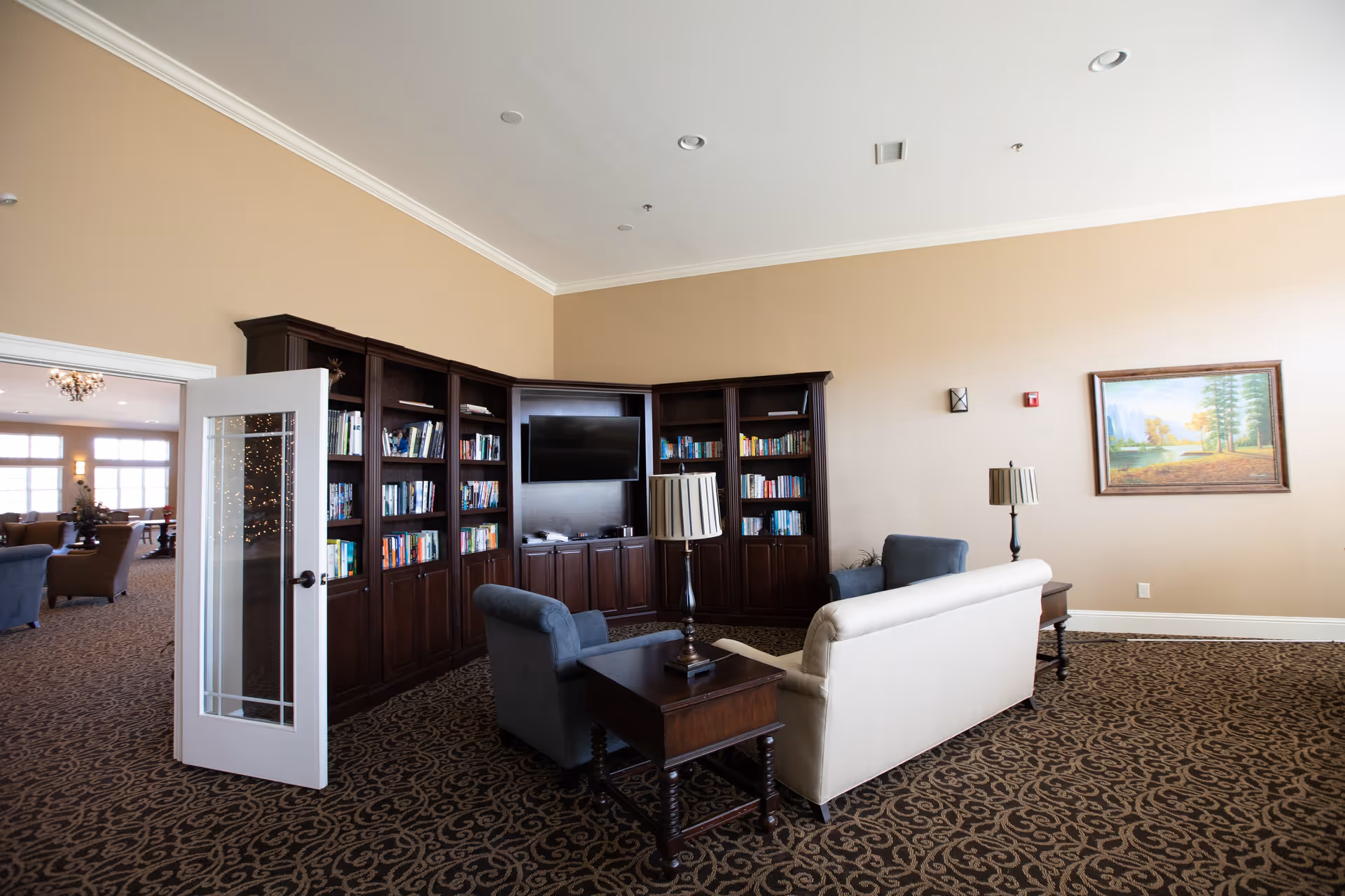 A cozy living room area in The Timberley featuring a beige sofa, two blue armchairs, a wooden side table with a lamp, and a large dark wood bookshelf with a mounted flat-screen TV. The room has beige walls, patterned carpet, and a framed landscape painting on the wall. A glass-paneled door opens to an adjacent room with more seating and large windows.