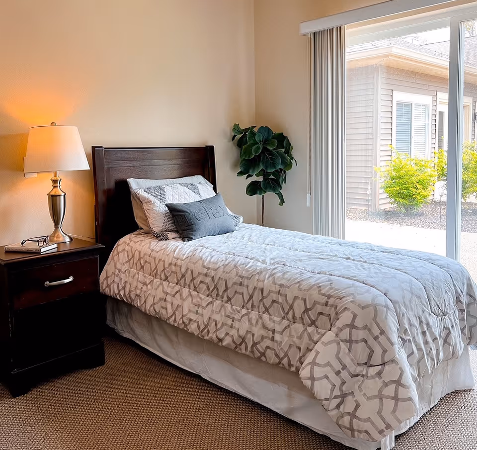 A neatly made single bed with patterned bedding and pillows in a bedroom. Next to the bed is a dark wooden nightstand with a lamp, a book, and a pair of glasses. There is a green potted plant in the corner and a large sliding glass door with vertical blinds that opens to an outdoor area with bushes and another building visible.