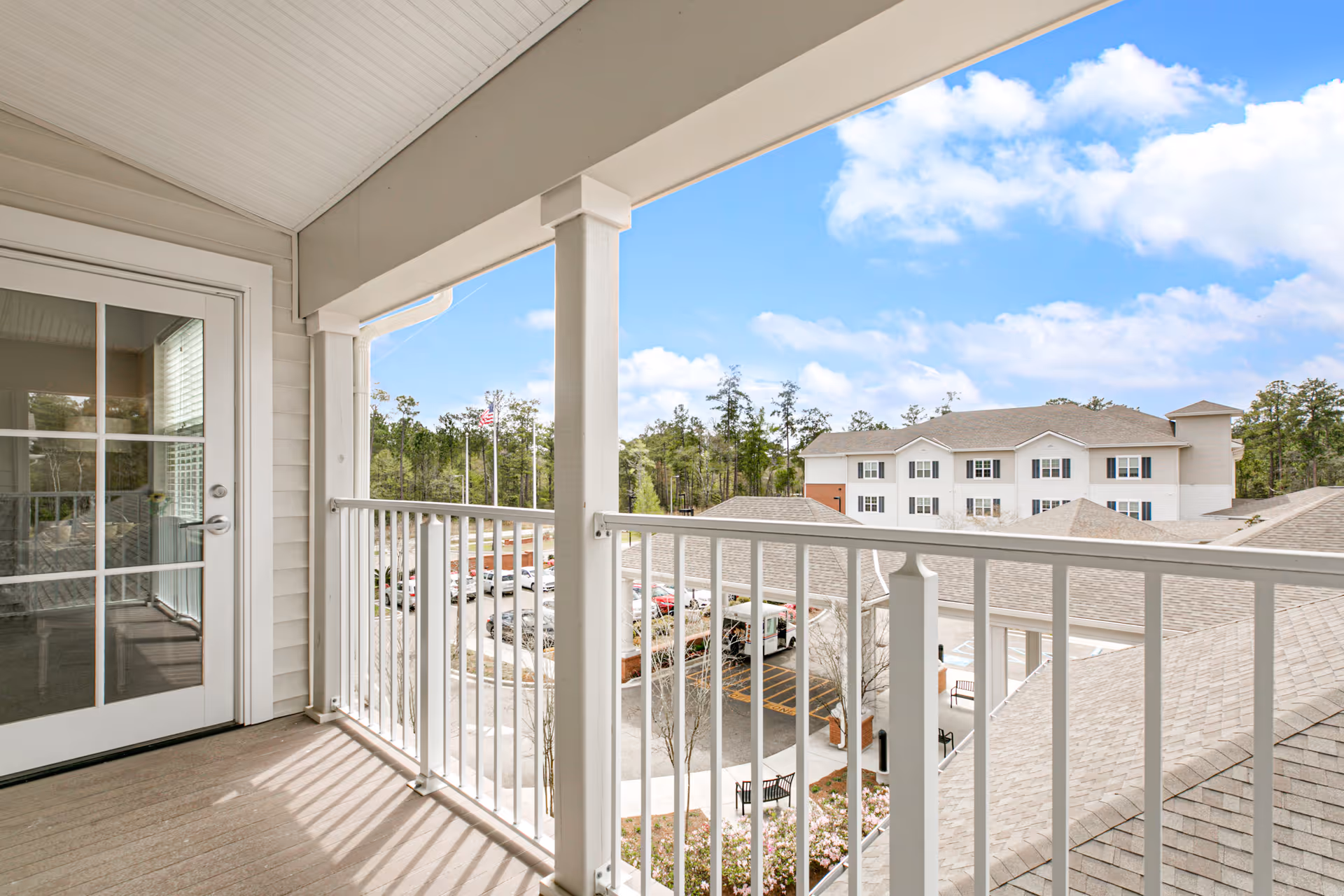 View from a covered balcony with white railing overlooking a parking area, landscaped garden, and a multi-story building with beige siding and multiple windows under a blue sky with scattered clouds.