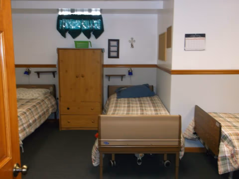 A small room with three single beds, each with plaid bedding. There is a wooden wardrobe against the back wall under a small window with a green valance. The walls are white with a wooden chair rail. A calendar and a cross are hung on the walls.