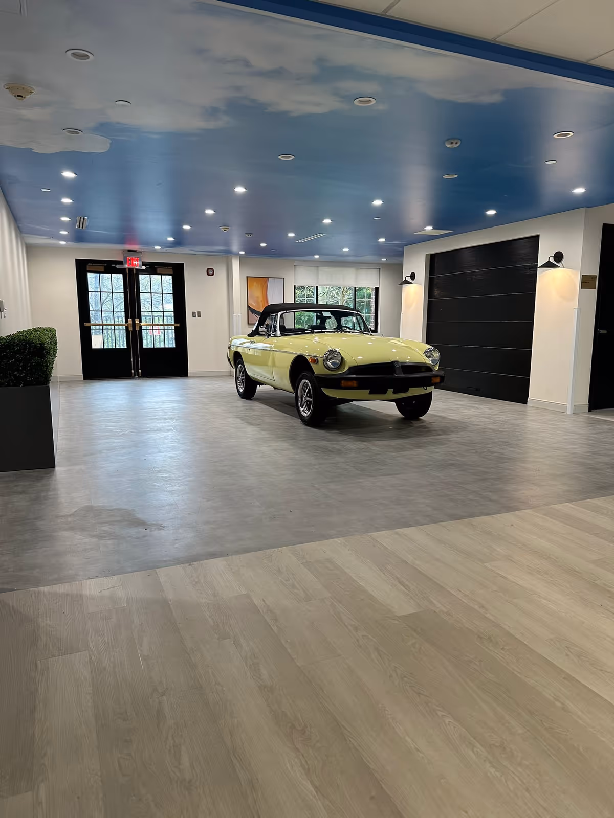 A spacious indoor area with a light yellow vintage convertible car parked in the center. The room has a blue ceiling with recessed lighting, light wood and gray flooring, large black double doors with glass panels, and a large window with a white blind. There is a black garage-style door on the right wall and a potted green plant on the left side.