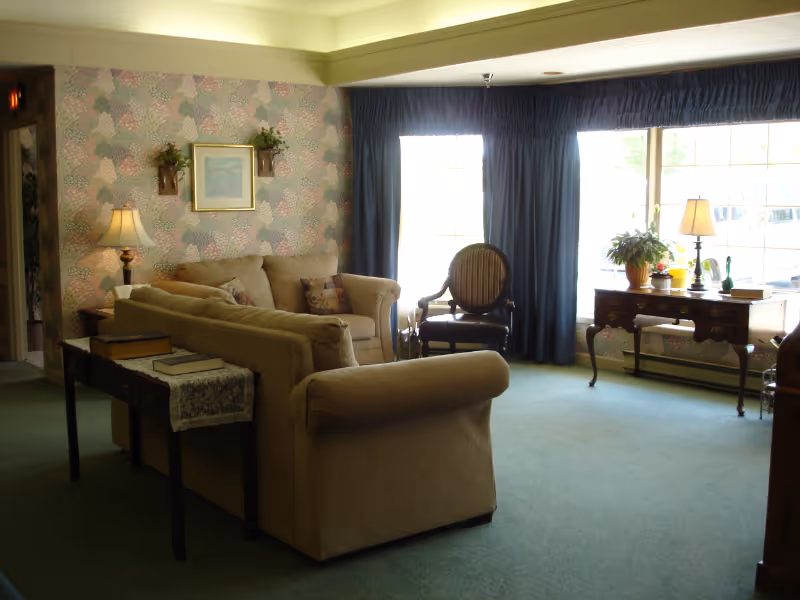 A well-lit common living room with upholstered sofas and chairs, side tables with lamps, and large curtained windows.