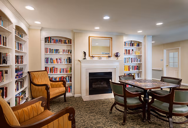 Cozy senior living library/lounge with built-in bookshelves flanking a fireplace, armchairs, and a table with chairs.