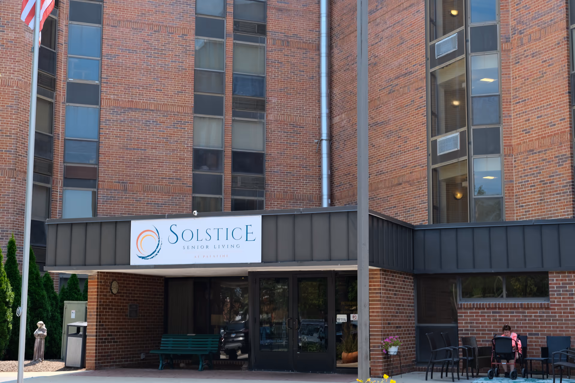 Entrance of Solstice Senior Living at Palatine, a multi-story brick building with large windows. There is a covered entryway with a sign displaying the facility's name. A green bench is placed under the covered area, and a person with a walker is seated at a table with chairs to the right of the entrance. An American flag is visible on the left side of the image.