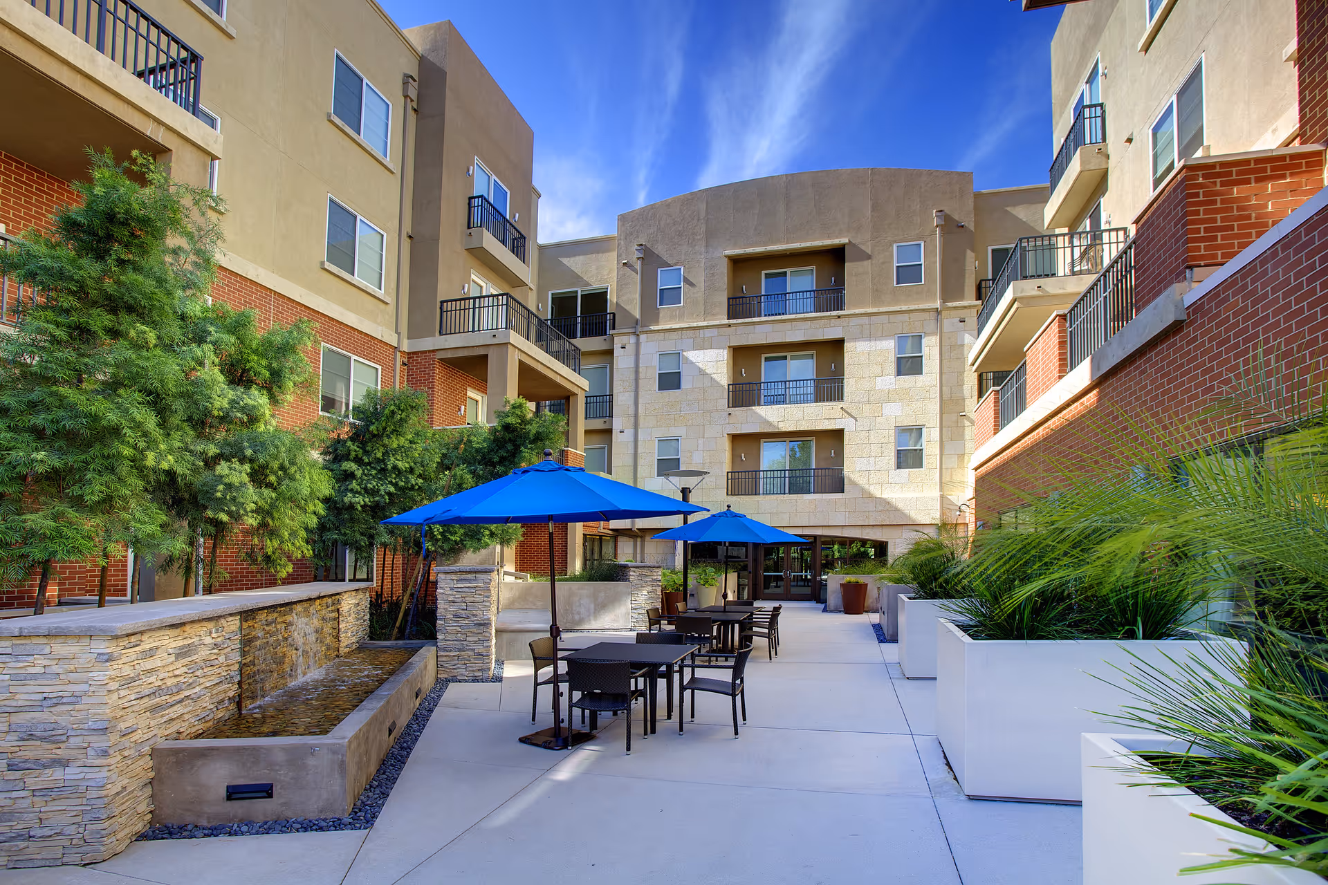 Outdoor courtyard area at Fountainview At Eisenberg Village featuring tables with blue umbrellas, chairs, a water feature with stone walls, and surrounding greenery. The courtyard is enclosed by a multi-story building with balconies and windows under a clear blue sky.