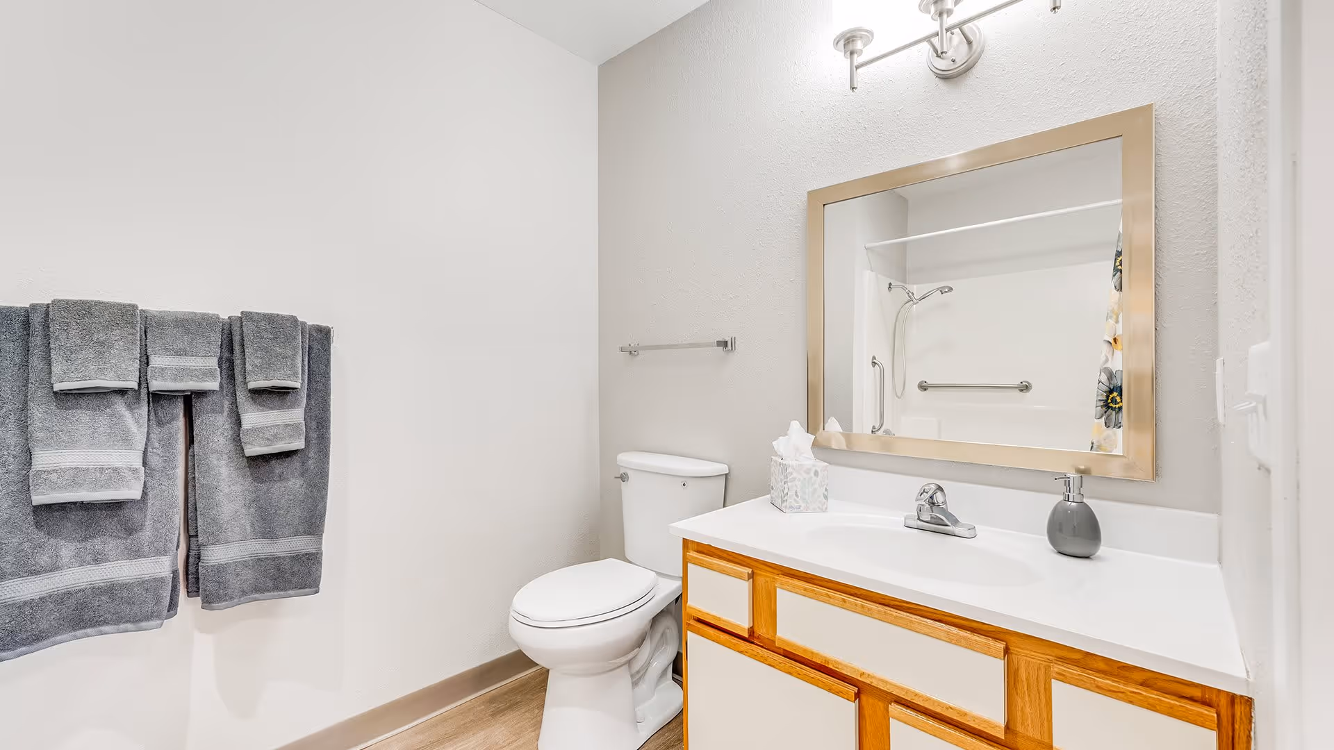 A clean and modern bathroom featuring a white toilet, a wooden vanity with a white countertop, a large rectangular mirror with a gold frame, a silver faucet, a soap dispenser, and a tissue box. Three gray towels hang on a towel rack on the left wall. The shower area with a grab bar and floral shower curtain is visible in the mirror's reflection.