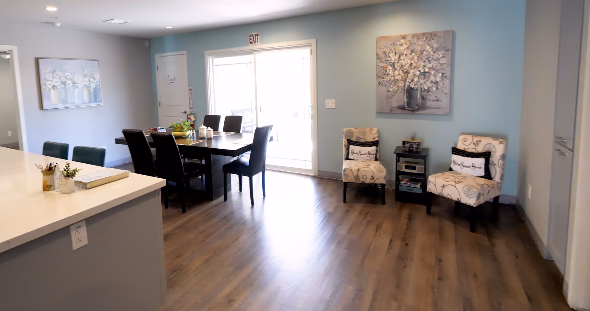 Interior view of a senior living facility dining and sitting area with a wooden dining table surrounded by six black chairs, two patterned armchairs with decorative pillows, a small side table with a stereo and books, and a kitchen counter with stools. The walls are painted light blue and gray, adorned with floral paintings, and there is a sliding glass door letting in natural light.