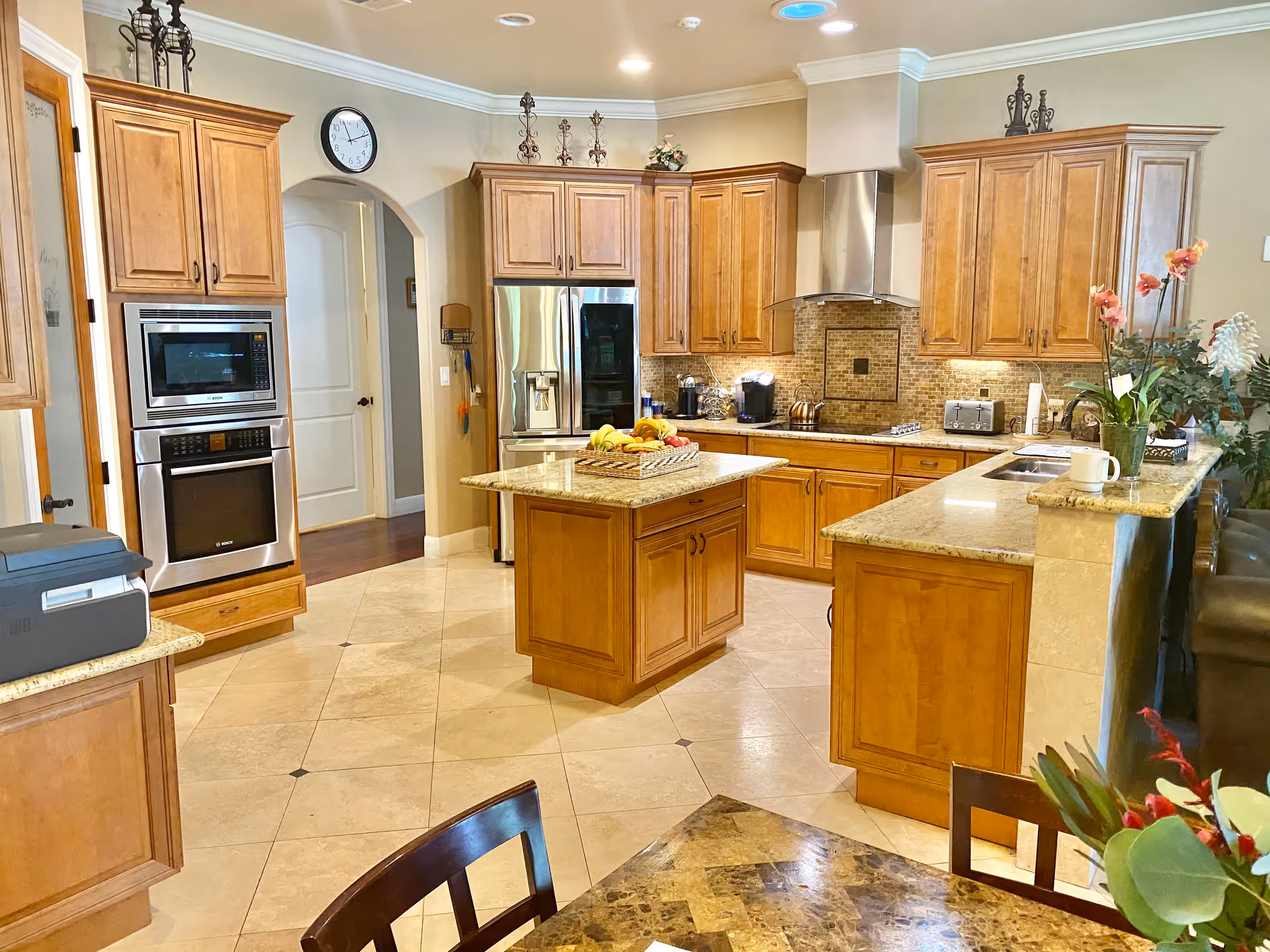 A spacious kitchen with wooden cabinets, granite countertops, and tiled floors. The kitchen features a central island with a basket of fruit, stainless steel appliances including a refrigerator, microwave, and oven. There is a stove with a stainless steel hood, a toaster, coffee maker, and a sink with a cup on the counter. Decorative plants and flowers are placed on the counters and a dining table with chairs is partially visible in the foreground.