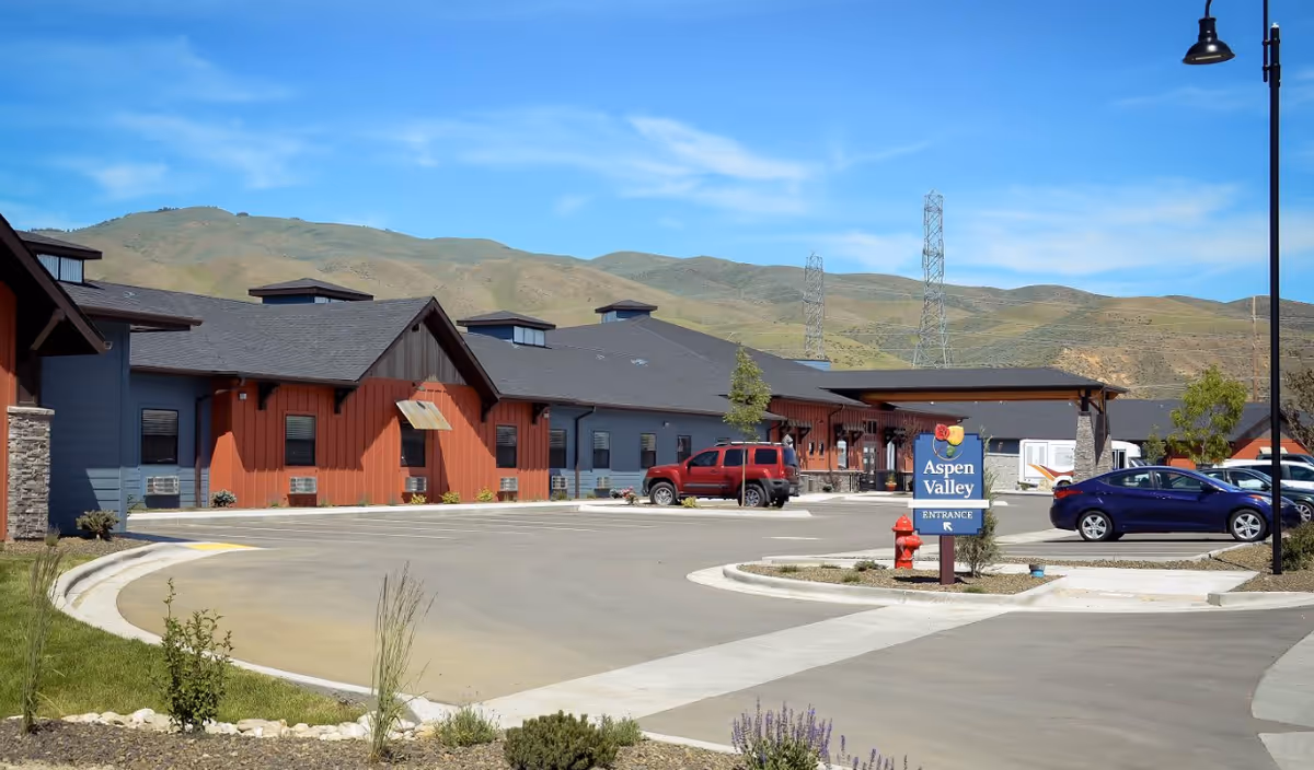 Front exterior of Aspen Valley Senior Living with a driveway, parking lot, entrance sign and mountains in the background.