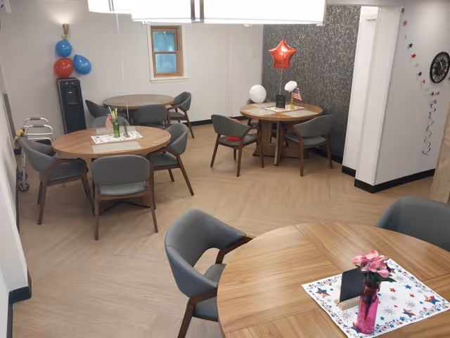 A small dining room with four round wooden tables, each surrounded by four gray cushioned chairs. The room has light wood flooring and white walls, with some patriotic decorations including balloons and small American flags on the tables. A walker is visible in the corner near a water dispenser.