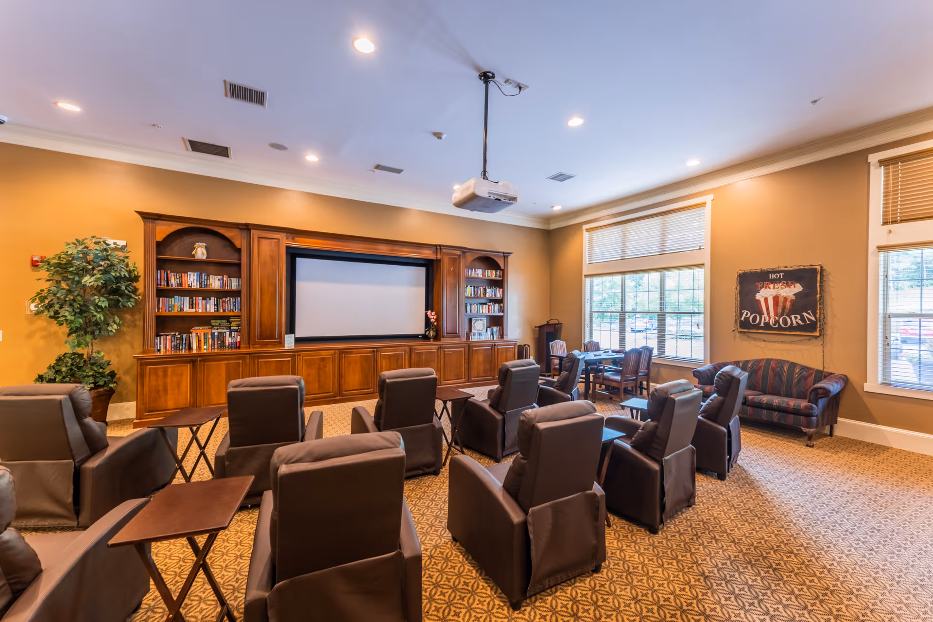 A cozy senior living common room with multiple brown recliner chairs arranged in rows facing a large screen mounted on a wooden entertainment center with bookshelves. The room has warm beige walls, large windows with blinds, a small table with chairs near the windows, a potted plant, and a vintage-style sign on the wall that reads 'Hot Fresh Popcorn'.