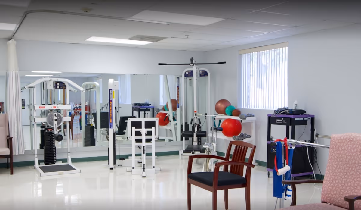 A senior living facility exercise room with various fitness equipment including weight machines, exercise balls, and a stationary bike. The room has white walls, a large mirror on one side, a window with vertical blinds, and several chairs including a wooden chair and a cushioned armchair.