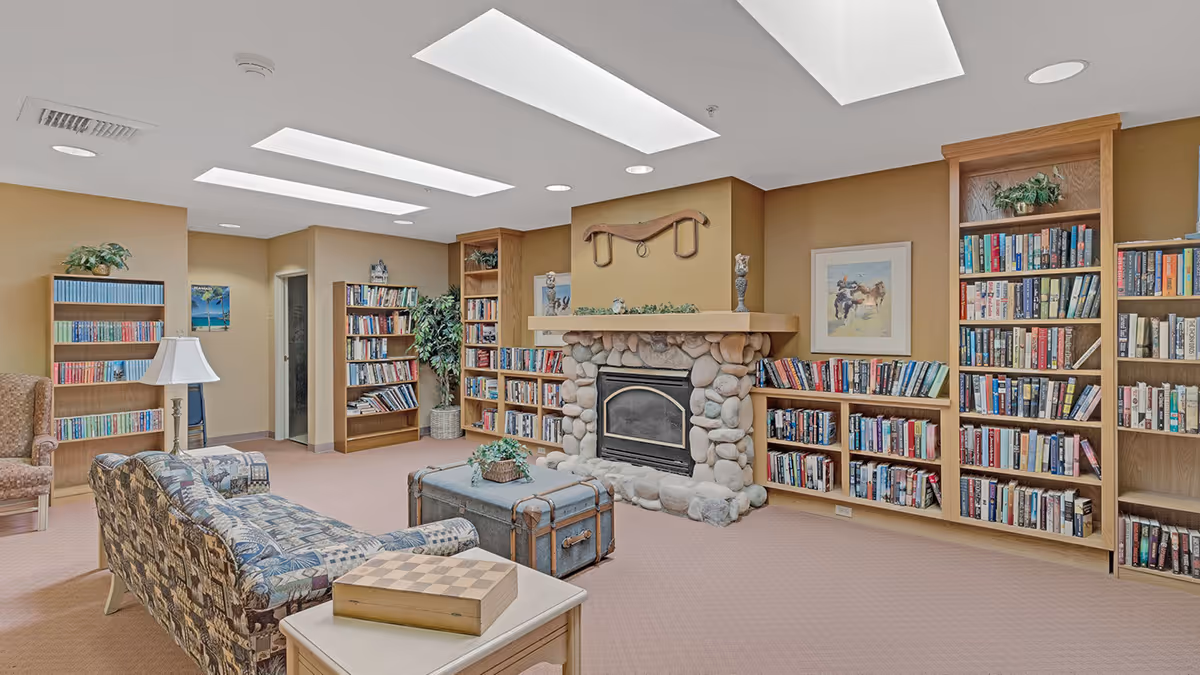A cozy library room with multiple wooden bookshelves filled with books, a stone fireplace in the center, a patterned sofa, an armchair, a trunk-style coffee table, a side table with a chessboard, and ceiling skylights providing natural light.