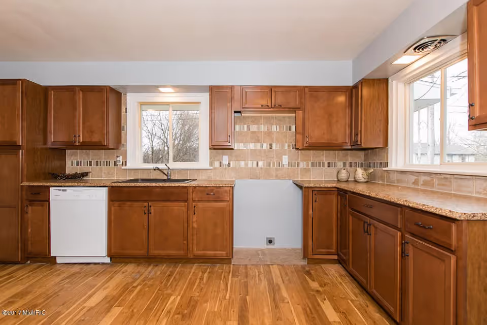 A kitchen with wooden cabinets and drawers, granite countertops, a white dishwasher, a double sink under a window, and a tiled backsplash. The floor is wooden, and there is a large window on the right side letting in natural light.