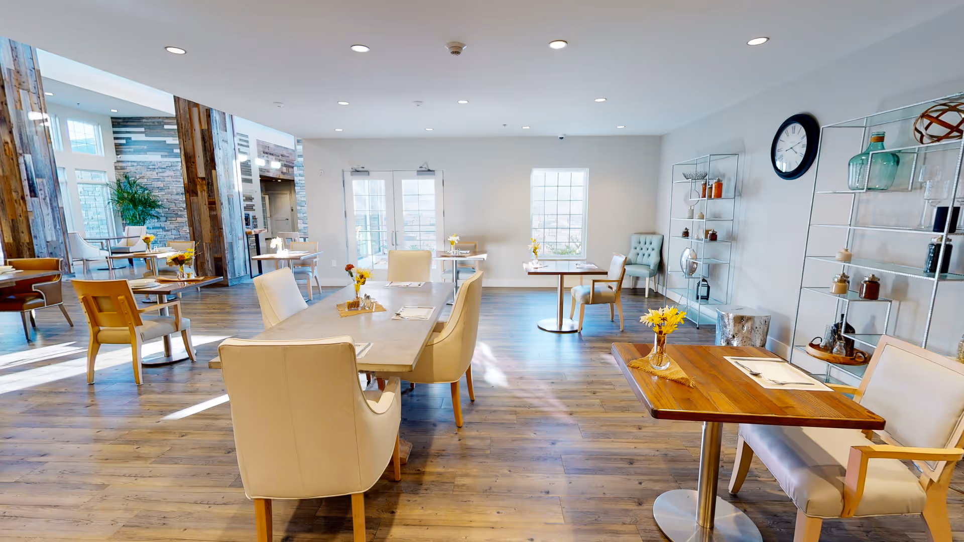 A bright and spacious dining room with wooden floors and modern furniture. Several tables are set with napkins and small vases of yellow flowers. The room has large windows letting in natural light, a clock on the wall, and decorative shelving with various items. The space has a clean, welcoming atmosphere.