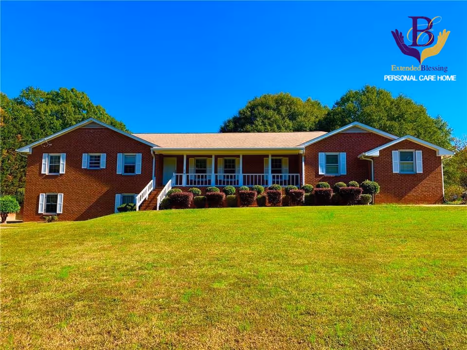 A single-story brick building with white window shutters and a front porch with white railings, surrounded by neatly trimmed bushes and a large grassy lawn under a clear blue sky.