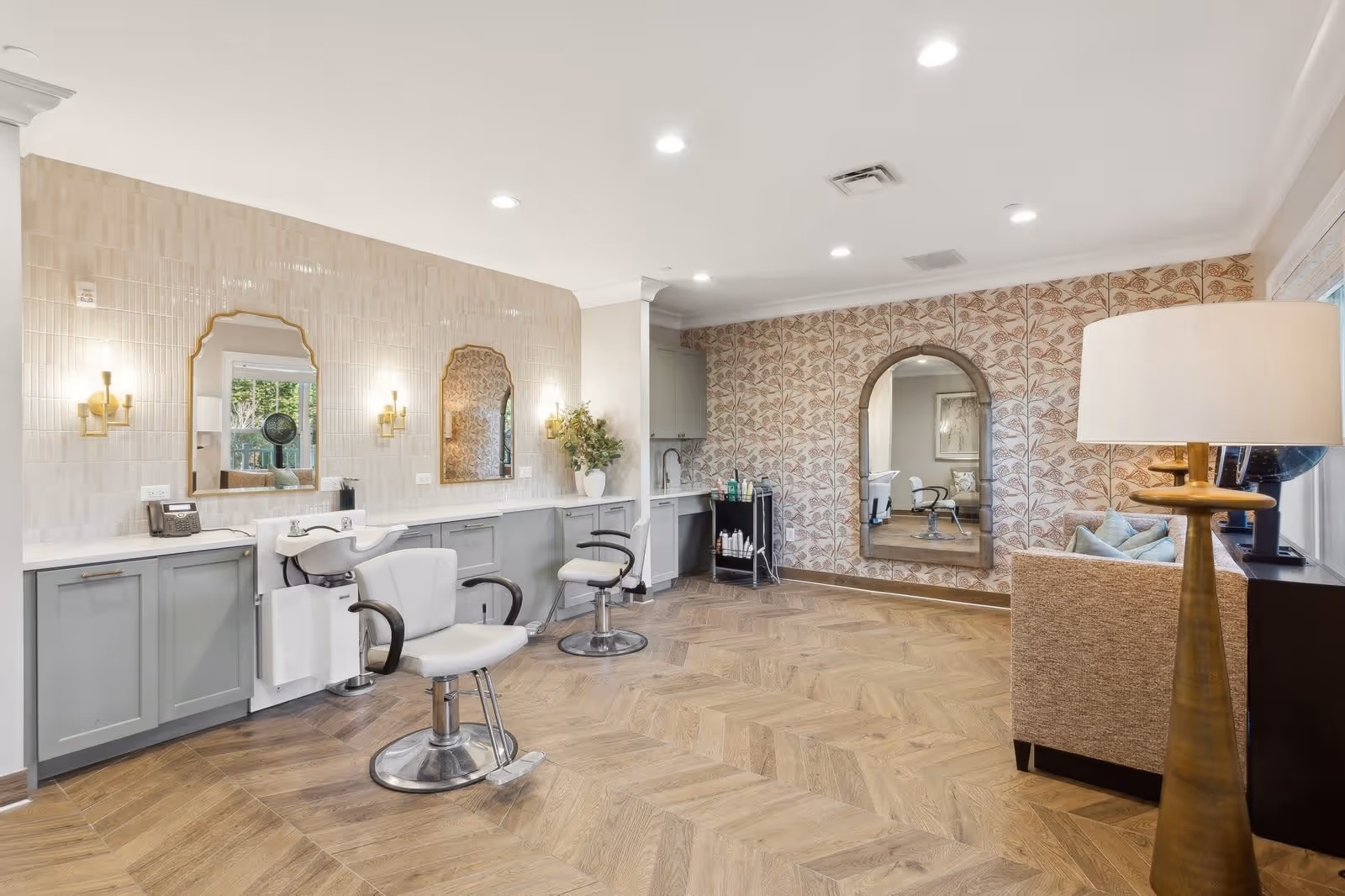 A bright and modern salon area in a senior living facility with two white salon chairs in front of mirrors mounted on a tiled wall. The room features wood-patterned flooring, a large decorative mirror on a wallpapered wall, a beige couch with blue cushions, and a tall floor lamp with a white shade.