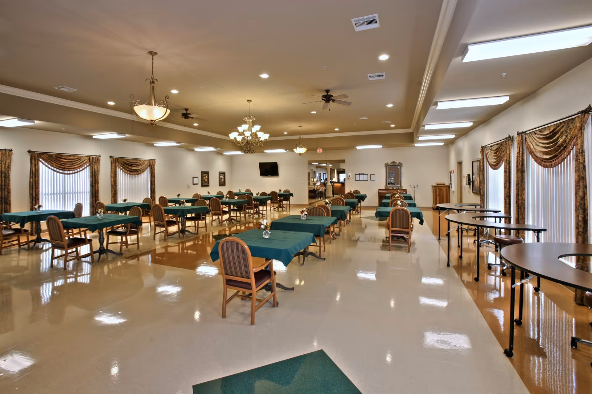A large dining room with multiple tables covered in green tablecloths and wooden chairs arranged neatly. The room has polished floors, several chandeliers and ceiling fans, large windows with patterned curtains, and a wall-mounted TV in the background.