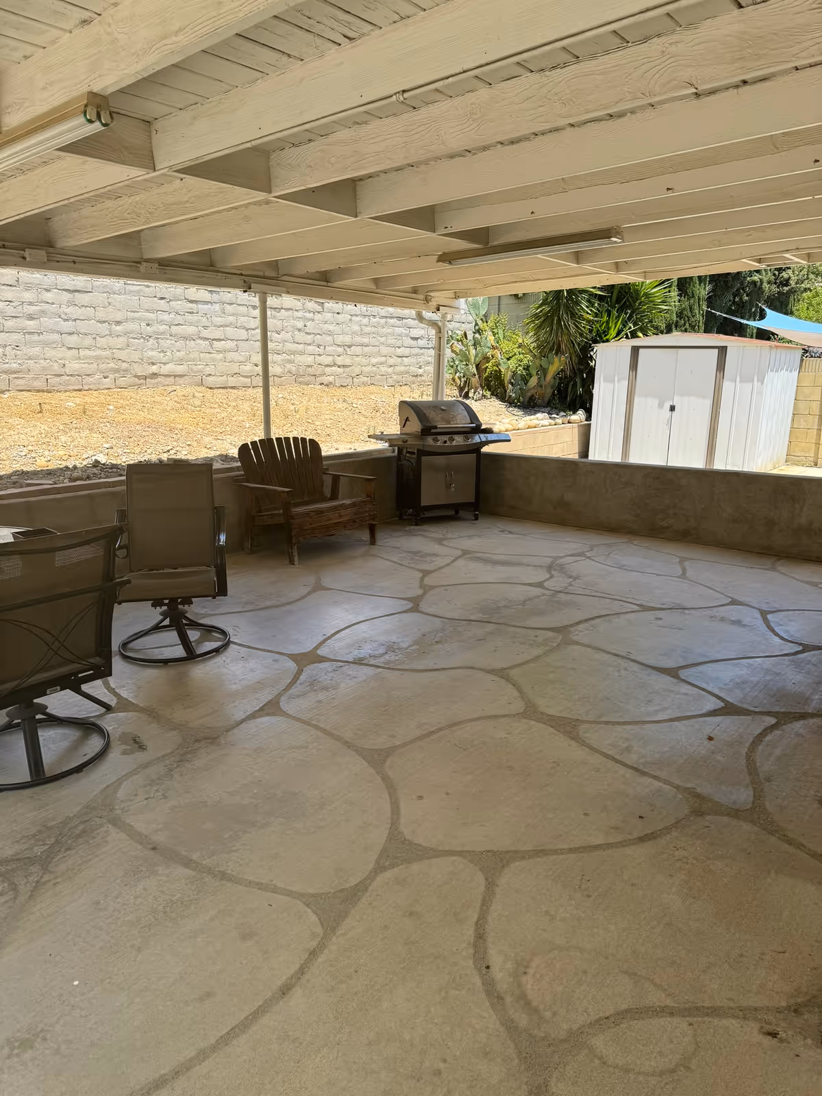 Covered outdoor patio with a stone-patterned concrete floor, patio chairs, a grill, and a small storage shed.