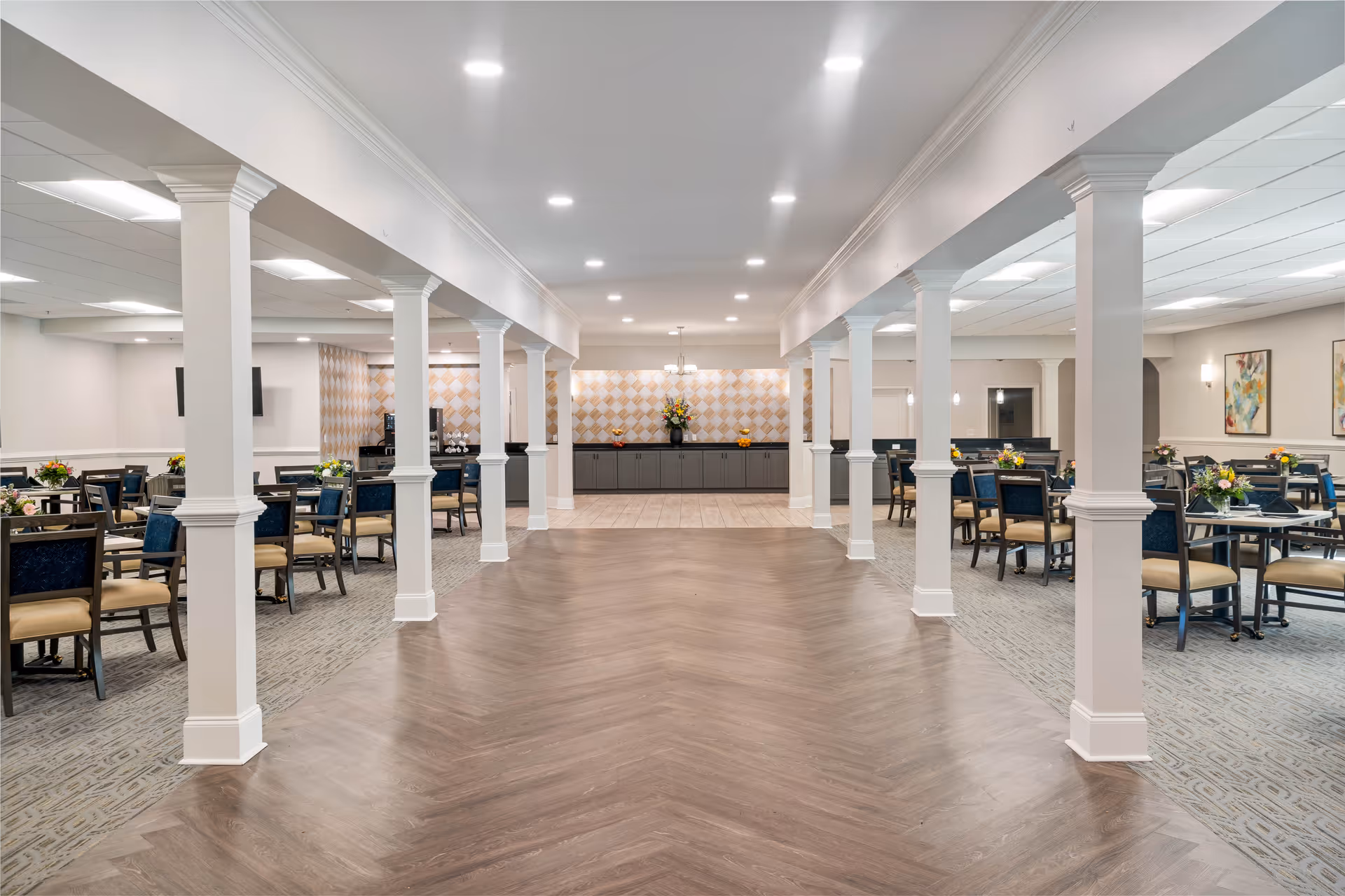 Large, well-lit communal dining room with columns, multiple dining tables and chairs, and a serving counter at the back.