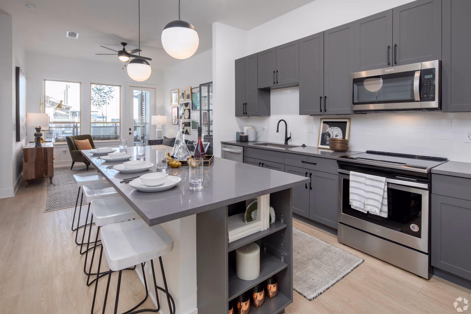 Modern kitchen and dining area with gray cabinets, stainless steel appliances including a microwave and oven, a large island with bar stools set with plates and glasses, pendant lights hanging above, and a living area with chairs and lamps in the background near large windows and a glass door.