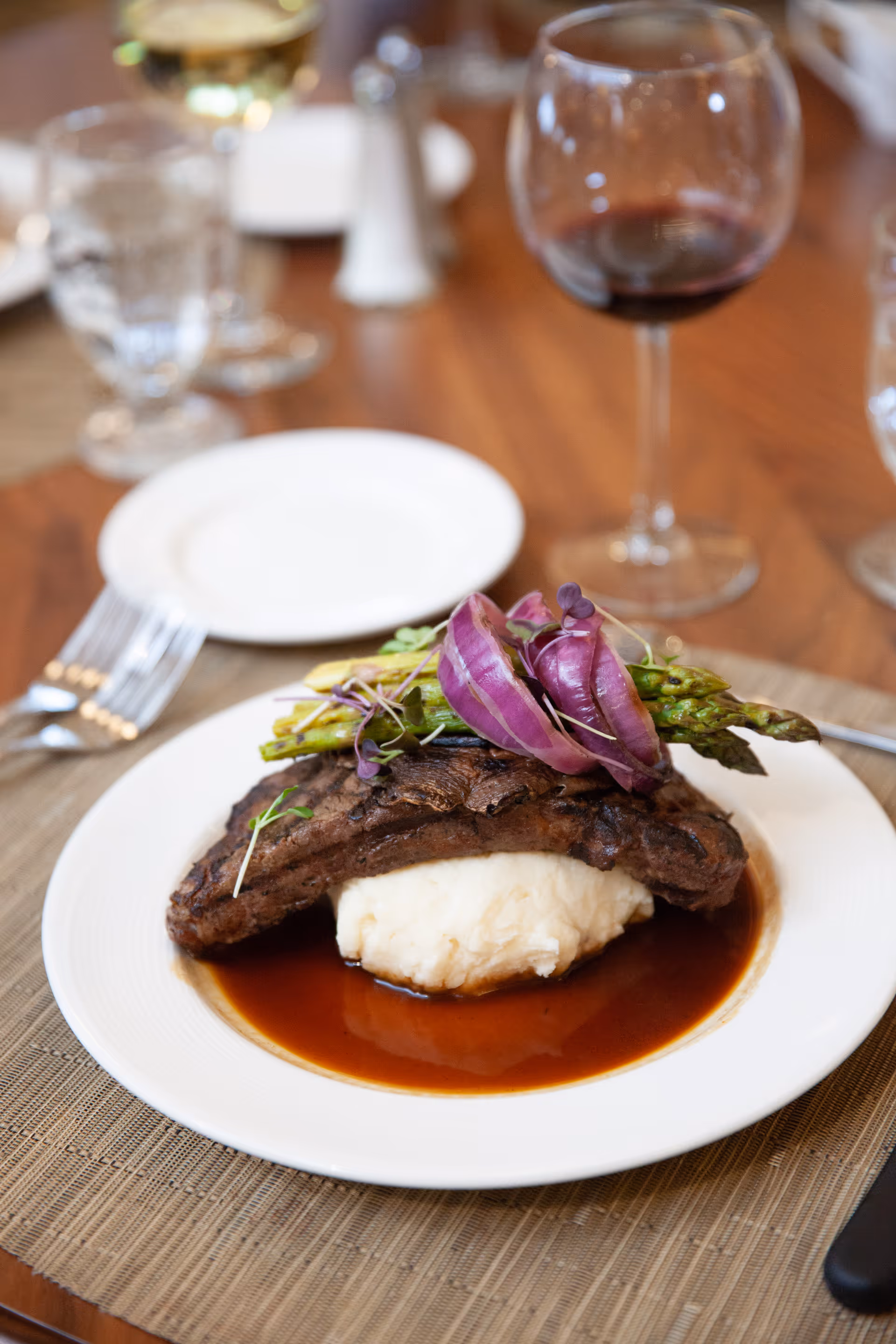 A plated steak on mashed potatoes topped with asparagus and red onions, set on a dining table with a wine glass in the background.