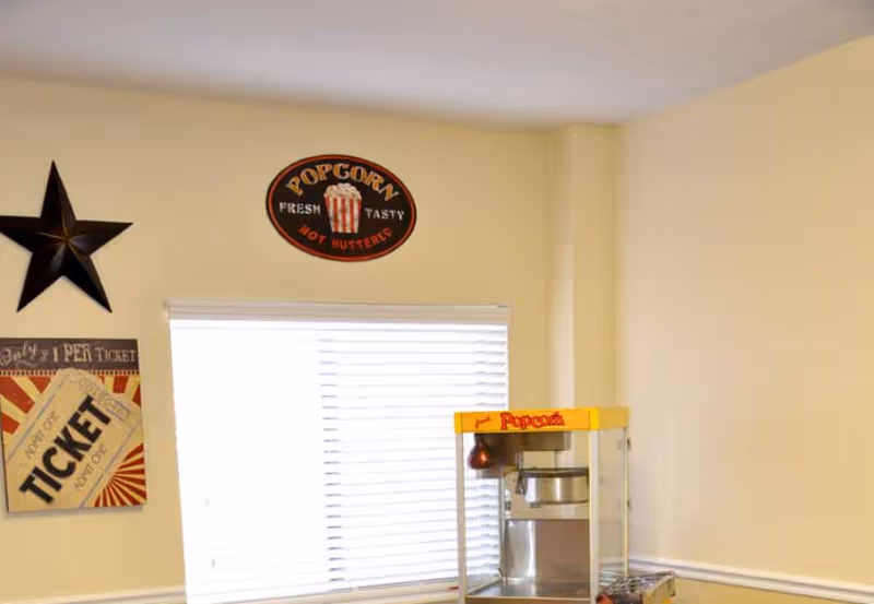 Interior corner of a room with a popcorn machine on a counter beneath a window with white blinds. The walls are light beige and decorated with a black star, a vintage-style popcorn sign, and a ticket-themed wall art.