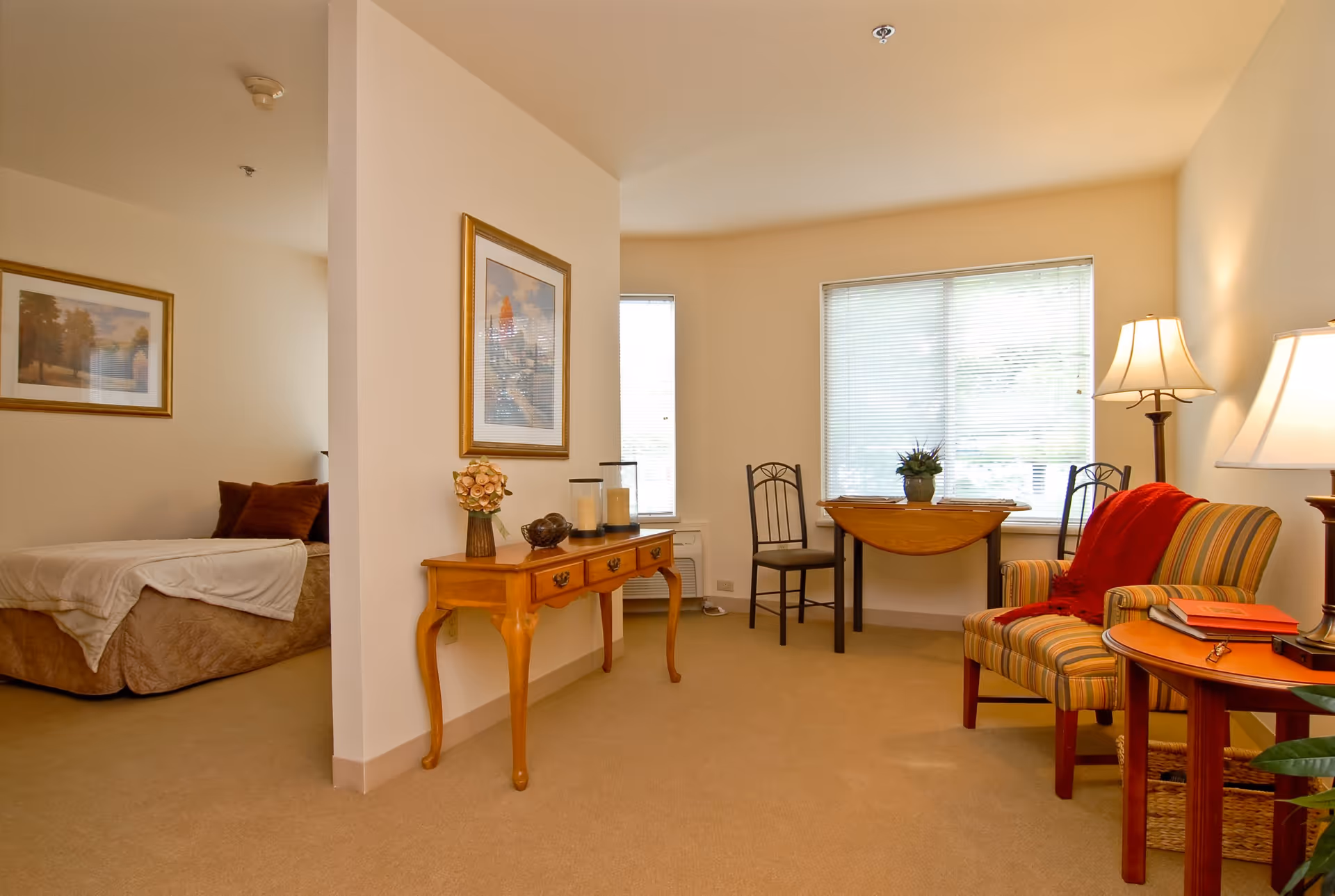 A cozy living space in a senior living facility with a small wooden table holding decorative items, a striped armchair with a red throw blanket, a side table with books and a lamp, and a small dining table with two chairs near a window with blinds. A bedroom with a bed and framed artwork is visible through an open doorway.