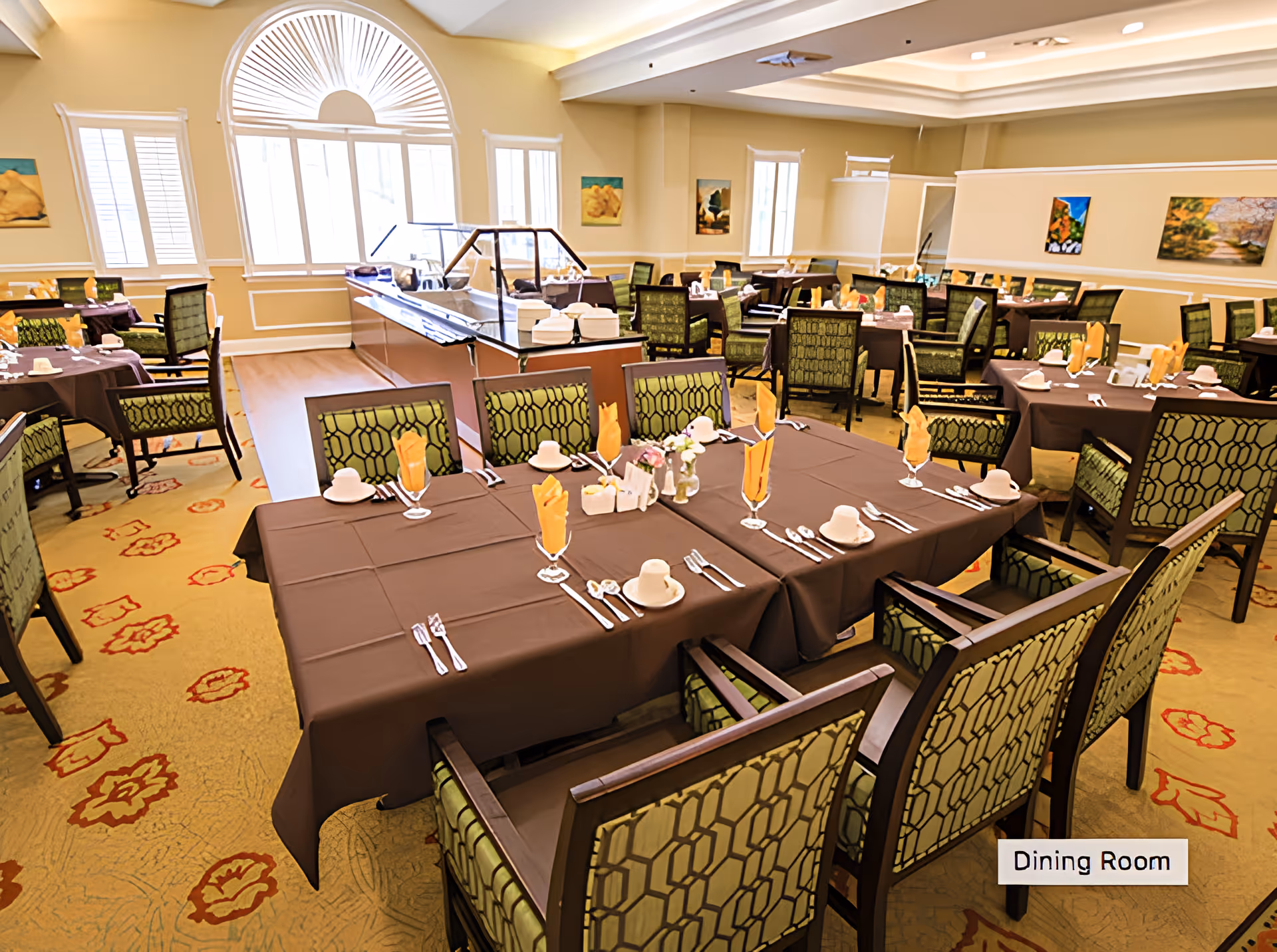 A spacious dining room with multiple tables covered in brown tablecloths, each set with utensils, napkins folded in glasses, and cups. The chairs have green patterned upholstery. Large windows with white blinds allow natural light to fill the room. The walls are decorated with framed artwork, and there is a buffet area in the center.