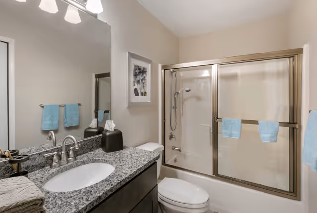A clean bathroom with a granite countertop sink, a large mirror, a toilet, and a bathtub with sliding glass doors. Light blue towels hang on the towel racks, and a framed picture is on the wall above the toilet.