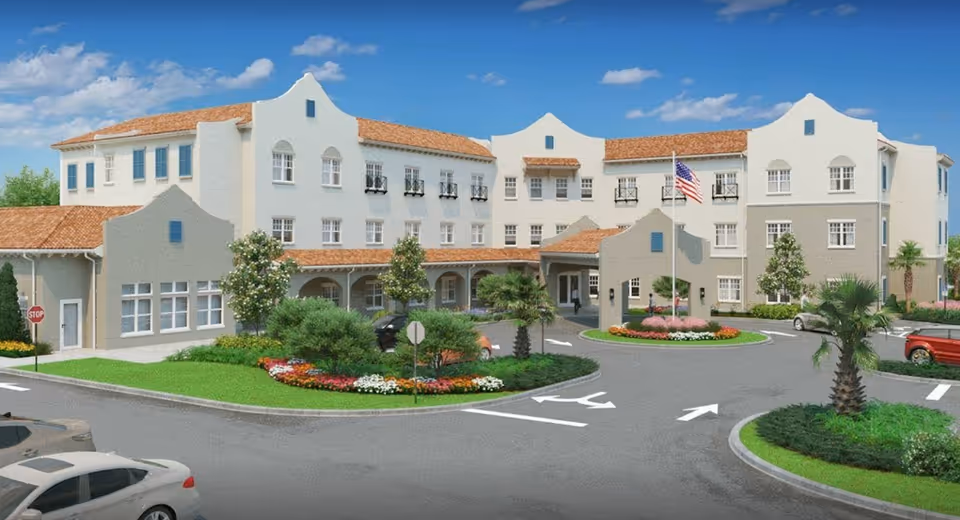 Front exterior of a three-story Mediterranean-style senior living building with a circular driveway, landscaped beds, parked cars, and an American flag.