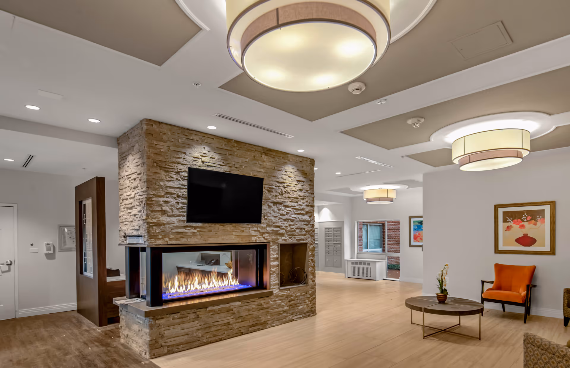Modern senior living lounge featuring a stone double-sided fireplace, wall-mounted TV, seating, and contemporary ceiling lights.