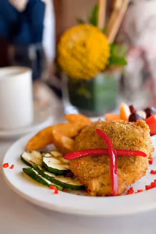 A close-up of a plate with breaded fried chicken garnished with two red pepper strips crossed on top, accompanied by grilled zucchini slices, potato wedges, and a small serving of mixed fruit. In the background, there is a white cup and a blurred yellow flower in a green vase.