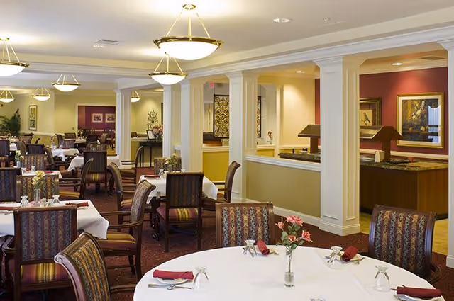 A spacious dining room in a senior living facility with multiple tables covered in white tablecloths, each set with glasses, napkins, and small flower vases. The room features patterned upholstered chairs, warm lighting from ceiling fixtures, and decorative artwork on the walls.